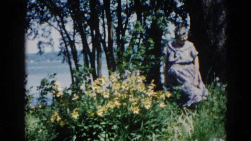 Woman in light dress among yellow wildflowers near a tree line, with what may be water visible in the background, Illinois circa 1962, 8mm.
