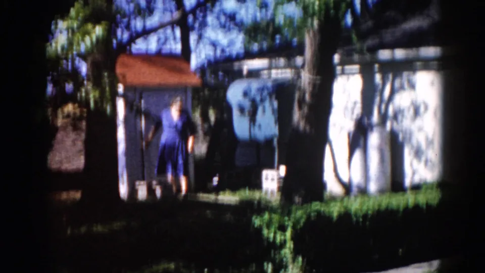 Woman in blue dress near a small outbuilding with reddish roof and white-painted wall, backyard scene from 8mm home movie, Illinois, circa 1962.