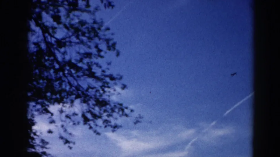 8mm home movie frame circa 1962, Illinois — tree-framed summer sky with cloud wisps and a faint jet contrail, with characteristic color shift of aged film.