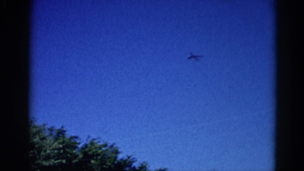 8mm home movie frame, circa 1962 Illinois: dark aircraft silhouette crossing a saturated blue sky with deciduous treetops at lower left.