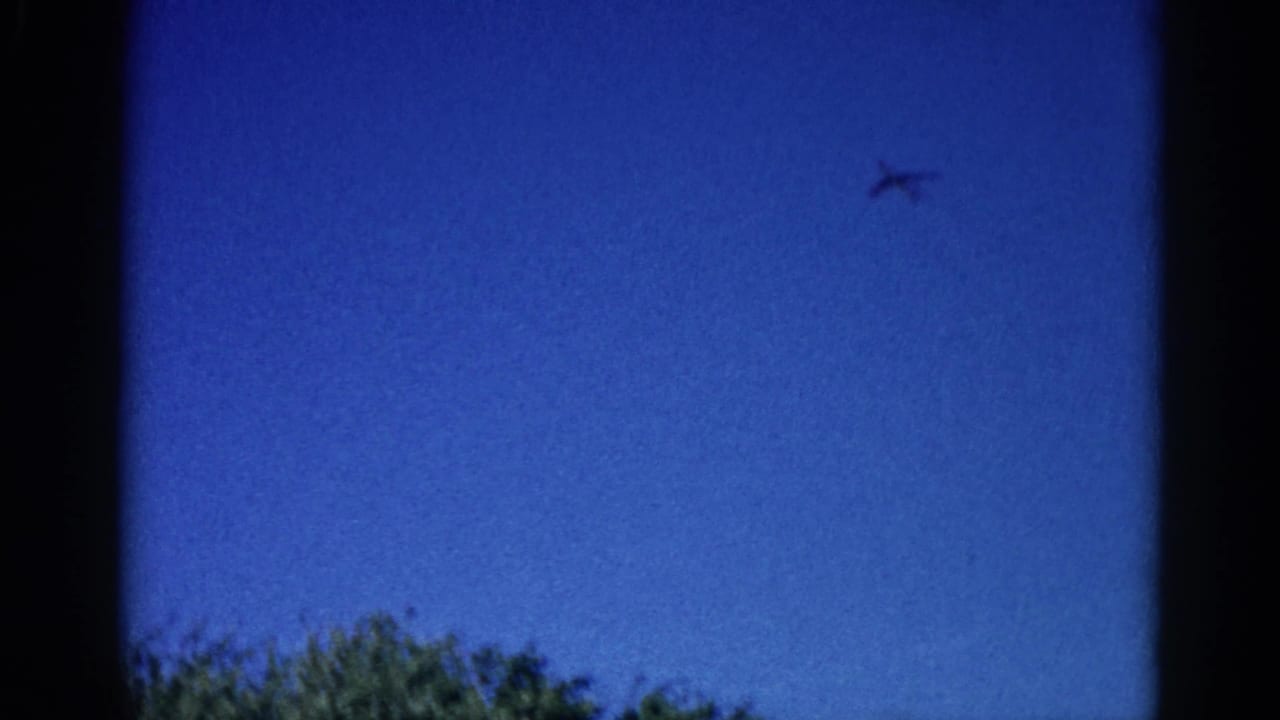 8mm home movie frame, circa 1962 Illinois: aircraft tracking right across saturated blue sky, deciduous treetops framing lower left edge.
