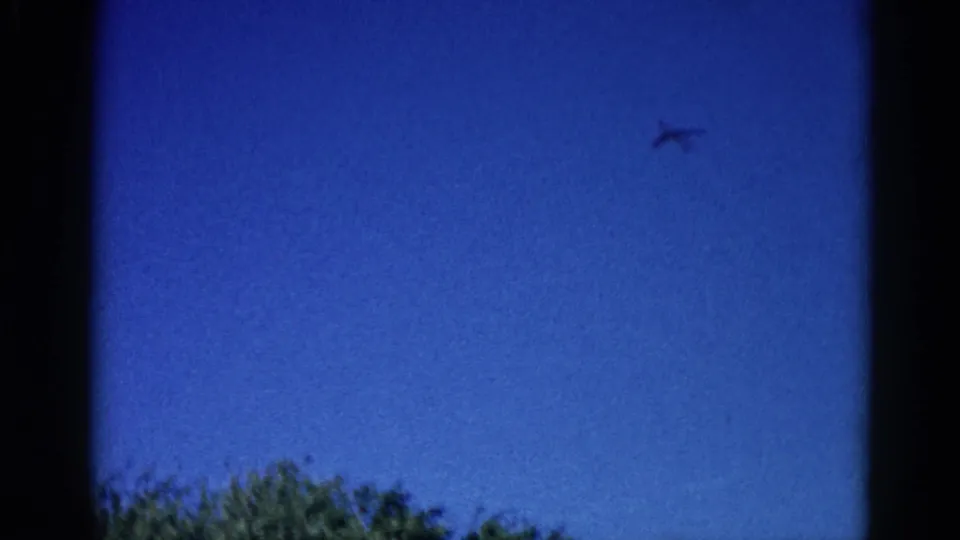 8mm home movie frame, circa 1962 Illinois: aircraft tracking right across saturated blue sky, deciduous treetops framing lower left edge.