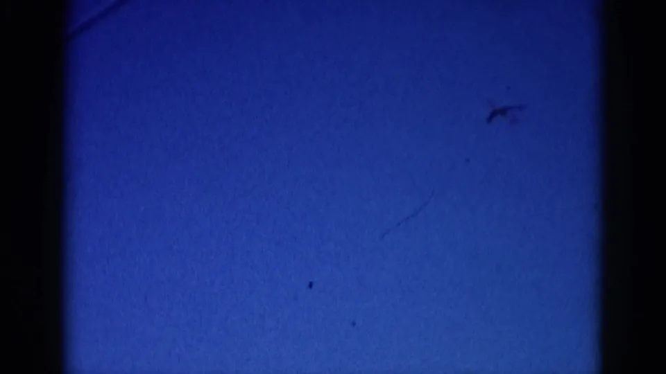 8mm home movie frame, circa 1962 Illinois: aircraft silhouette in mid-sky with a faint diagonal contrail forming below it, no tree canopy in frame.