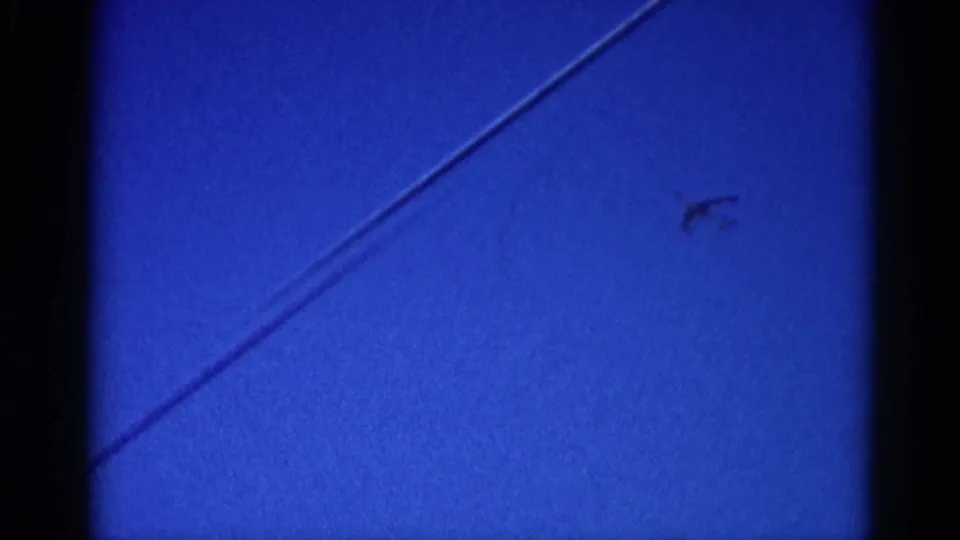 8mm home movie frame, circa 1962 Illinois: a bright diagonal vapor trail dominates the saturated blue sky with a small aircraft silhouette near its leading tip.