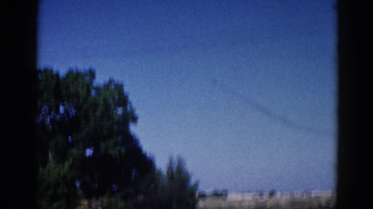 Opening 8mm home-movie frame from Illinois, c. 1962 — blue sky, leafy deciduous trees, diagonal power lines, and a warm horizon band, shot through a window.