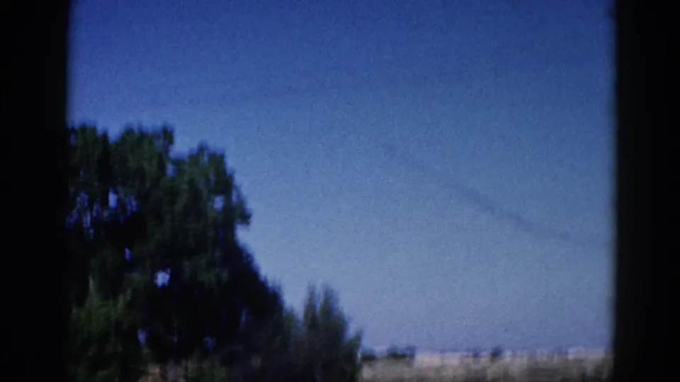 Opening 8mm home-movie frame from Illinois, c. 1962 — blue sky, leafy deciduous trees, diagonal power lines, and a warm horizon band, shot through a window.