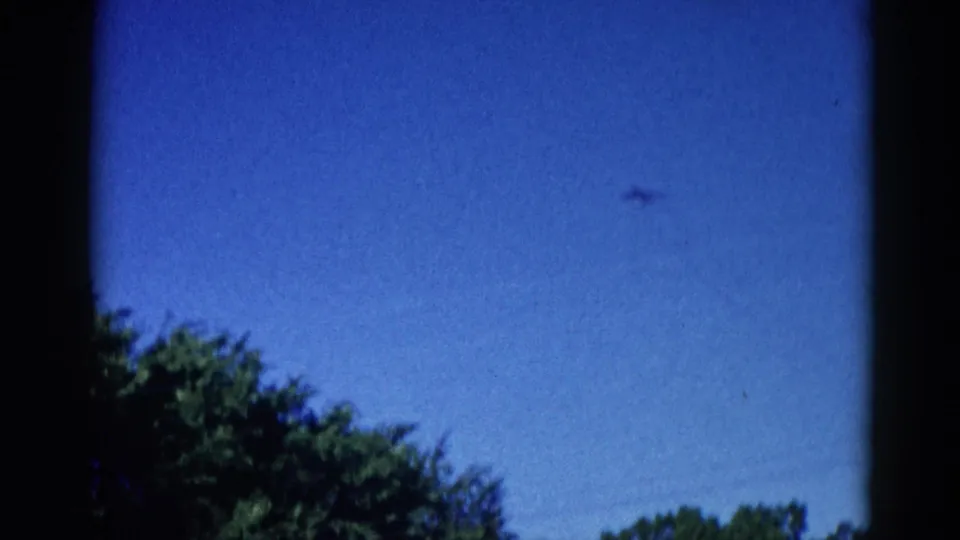 Mid-sequence 8mm frame from Illinois, c. 1962 — small aircraft crossing a deep blue sky above tree canopy, viewed through a window.