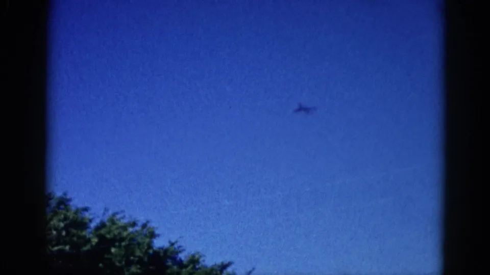 Closing 8mm frame from Illinois, c. 1962 — aircraft now left of center against deep blue sky, tree canopy at bottom, window border surrounds.