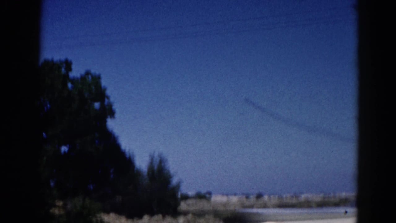 8mm home movie frame, Illinois 1962: faint contrail arcing across a clear blue sky above a flat rural landscape, framed by window edges and leafy trees at left.