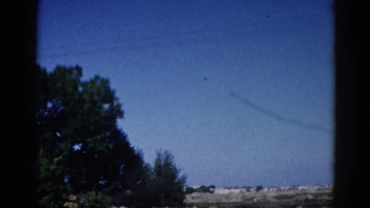 8mm home movie frame, Illinois 1962: faint contrail arcing across blue sky above flat Midwest landscape, large deciduous tree at right, window border visible.