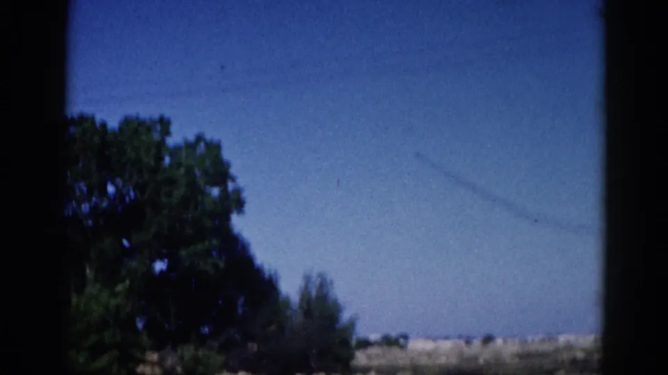 Mid-sequence 8mm home movie frame, Illinois 1962: faint contrail crossing a blue sky above a flat rural horizon, leafy trees at lower left, window edges visible.