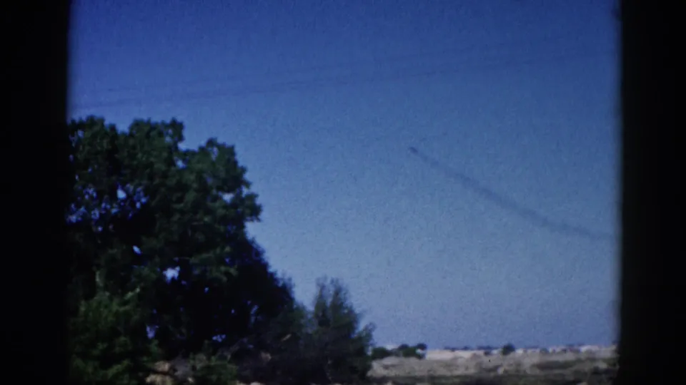 Final 8mm home movie frame, Illinois 1962: faint contrail in blue sky above flat rural landscape, large deciduous tree at right, window border visible.