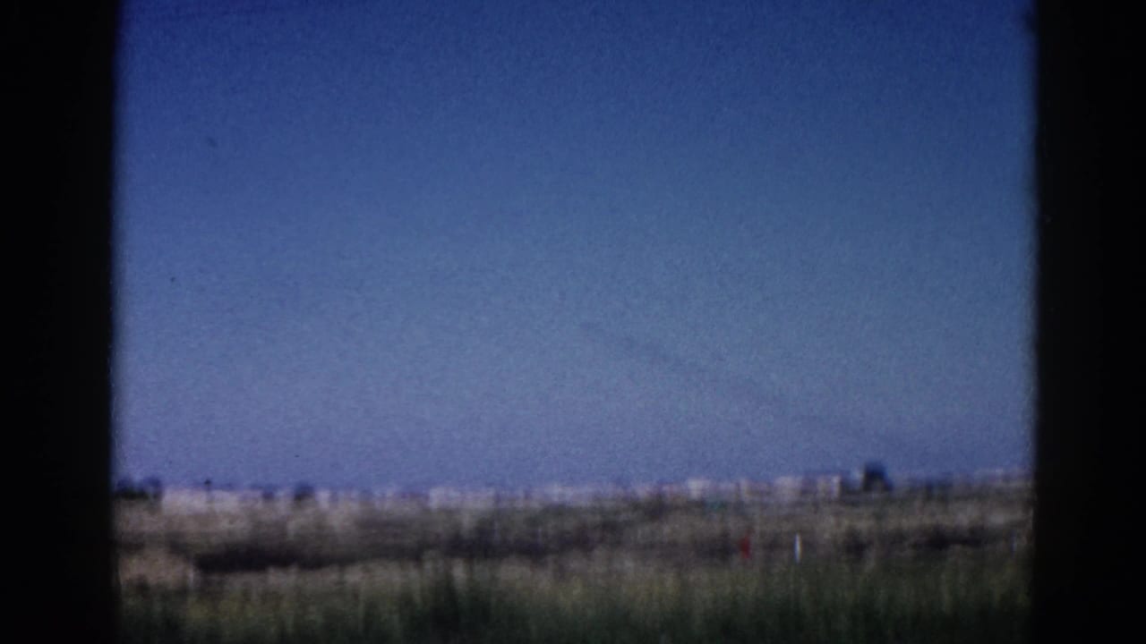 8mm home movie, 1962 Illinois — wide prairie view through car window, contrail across the sky, low indistinct structures on the far horizon