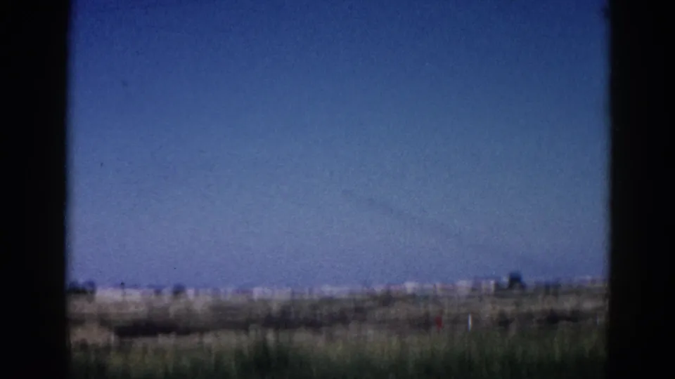8mm home movie, 1962 Illinois — wide prairie view through car window, contrail across the sky, low indistinct structures on the far horizon