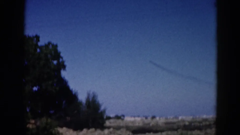 8mm home movie, 1962 Illinois — silhouetted tree enters the right of frame as the vehicle passes, open blue sky above flat rural land