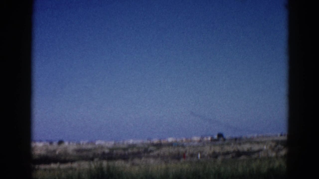 Flat open landscape viewed through dark frame edges, with clear blue sky and a faint diagonal contrail, 8mm film.