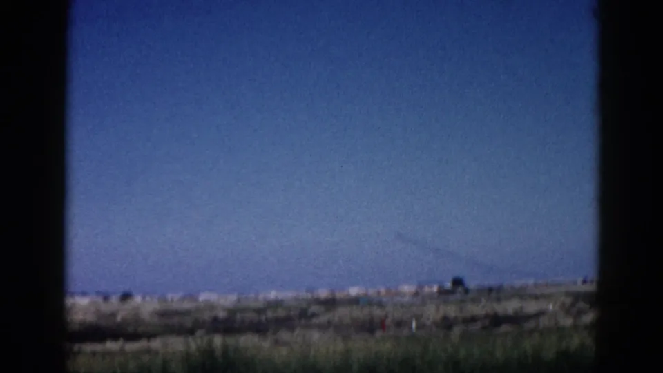 8mm home movie, Illinois c.1962; flat grassy terrain in foreground, vast blue sky with a faint contrail crossing diagonally, dark window or door edges framing the shot.