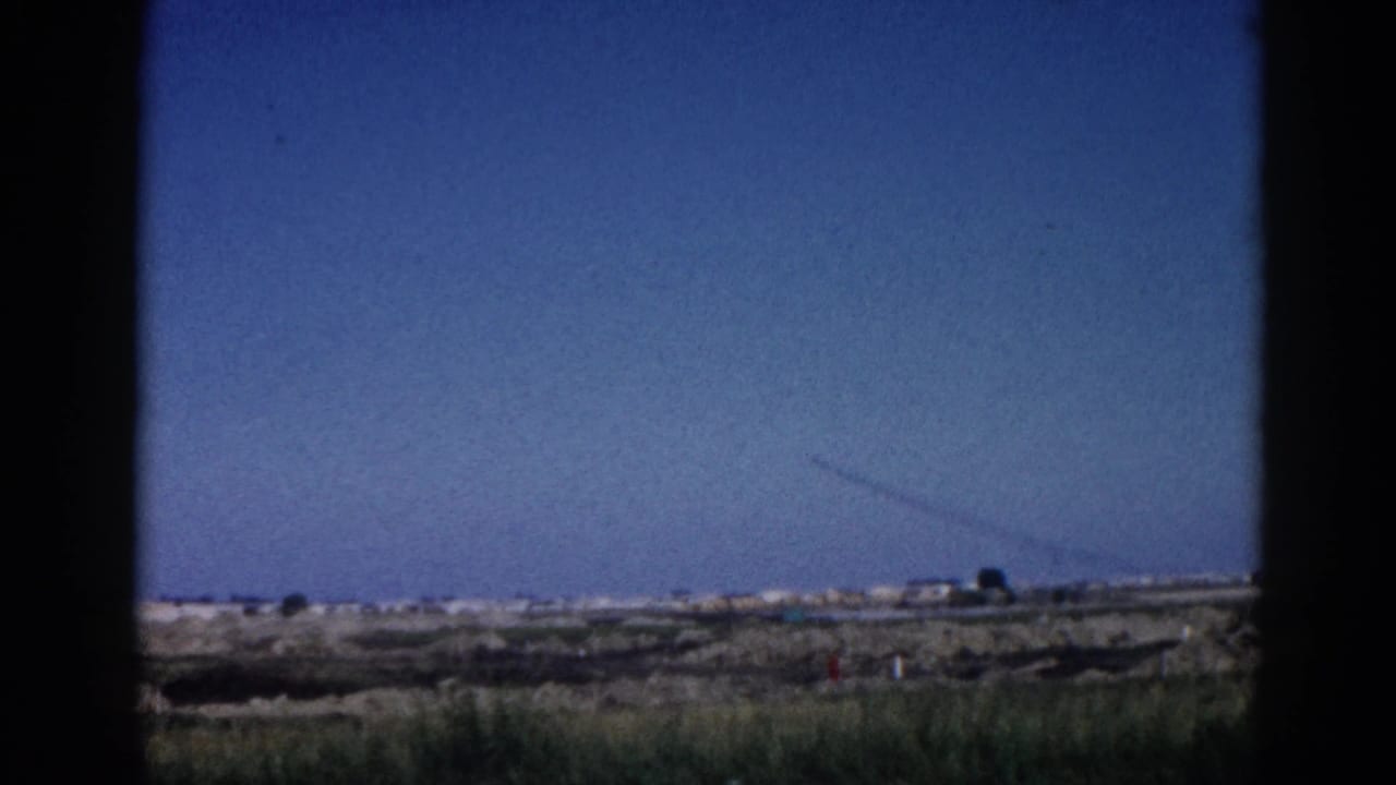 Wide flat landscape under clear blue sky; a diagonal contrail streaks across the upper sky, dark right edge frames the view, 8mm film.