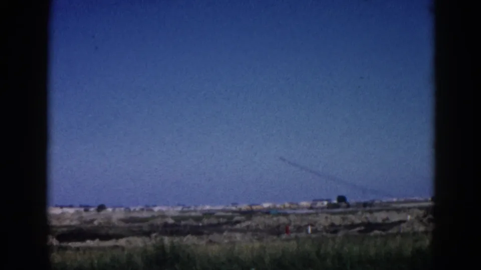 8mm home movie, likely Illinois c.1962; expansive blue sky with a contrail, flat scrubby terrain on the horizon, framed by dark vertical edge suggesting a window or vehicle opening.