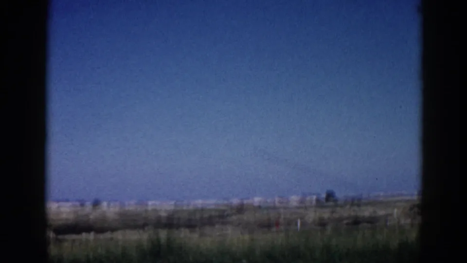 8mm home movie, c.1962 Illinois; sky fills most of the frame, a faint contrail traces across the blue, sparse low vegetation visible along the bottom edge.