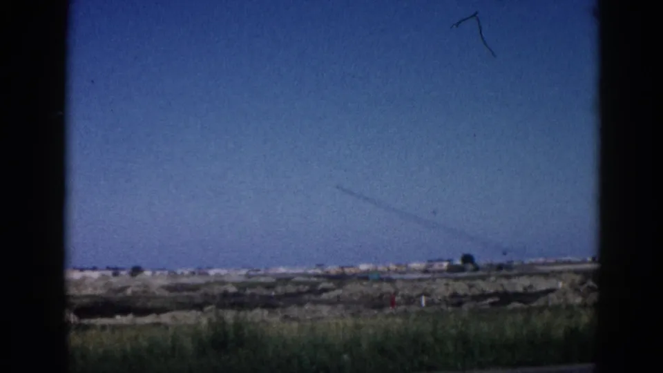 8mm home movie, c.1962 Illinois; open flat terrain at base of frame, blue sky with a contrail in the upper area, dark vertical framing edge at right suggests a window or vehicle.