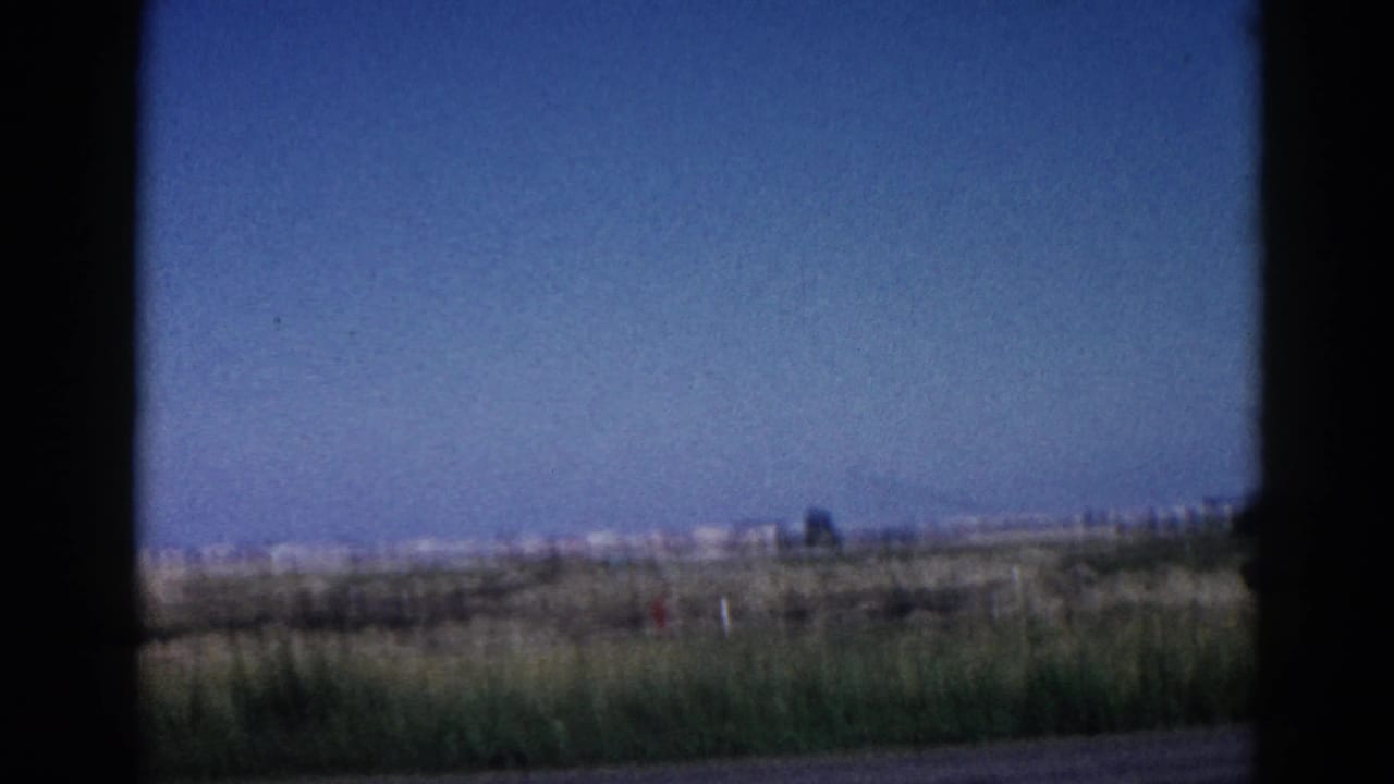 8mm home movie frame, c.1962 Illinois — flat rural landscape shot from a moving vehicle window, open fields and blue sky with distant horizon structures.