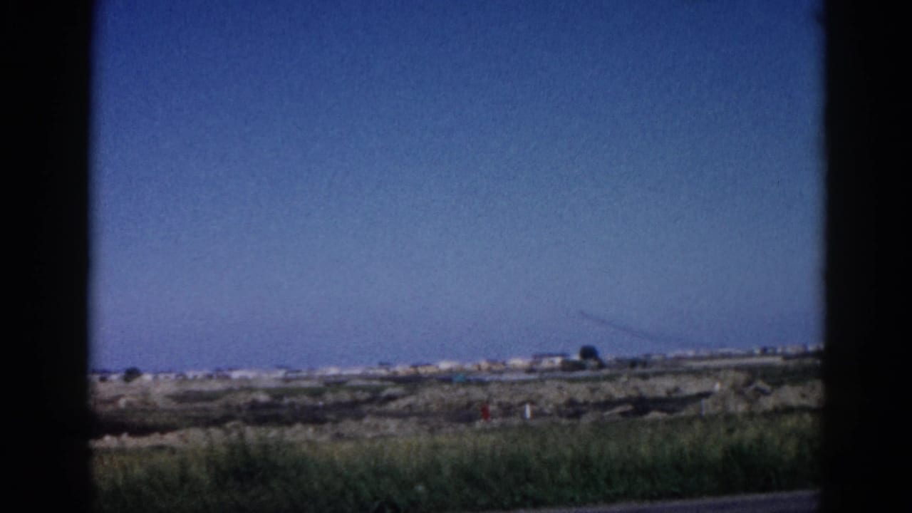 Vehicle window view of flat rural landscape with distant buildings and a tall vertical structure on the horizon under blue sky.