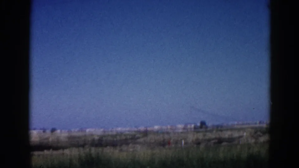 8mm home movie frame, c.1962 Illinois — sky-heavy window shot from a moving vehicle, distant crane or tower silhouette above flat open fields.
