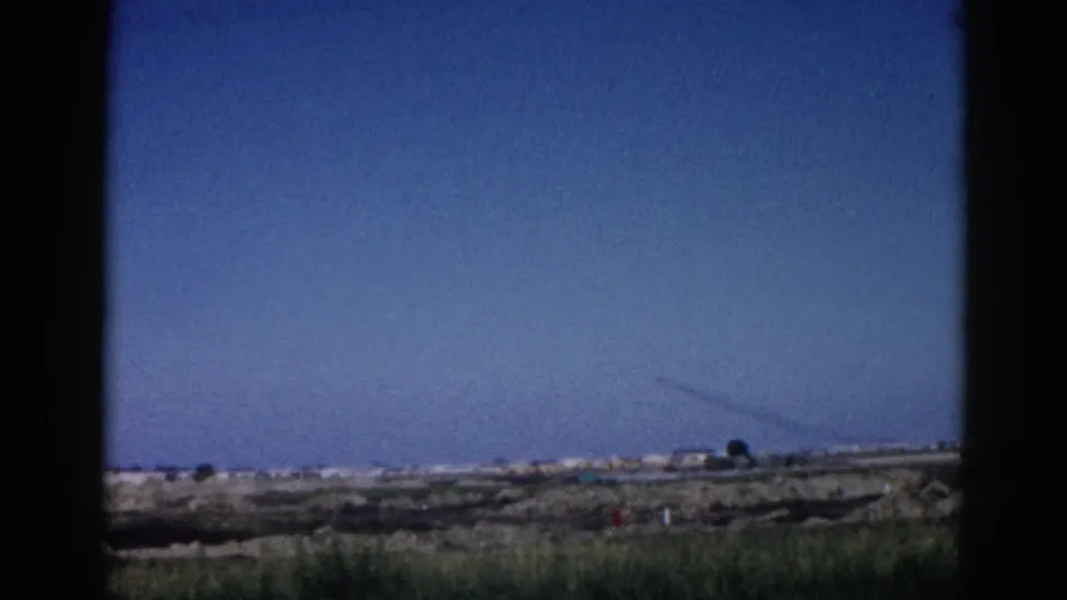 8mm home movie frame, c.1962 Illinois — final travel shot showing rural roadside landscape with distant structures and a crane-like silhouette.