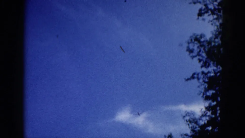 Tree silhouettes at frame edges with aircraft as small diagonal mark in blue sky, 8mm footage circa 1962.