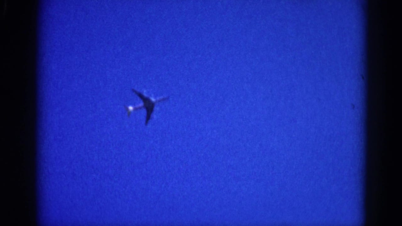 8mm home movie frame from 1962 Illinois showing a commercial jet as a dark silhouette against a uniformly deep blue sky, with a bright light near the nose.
