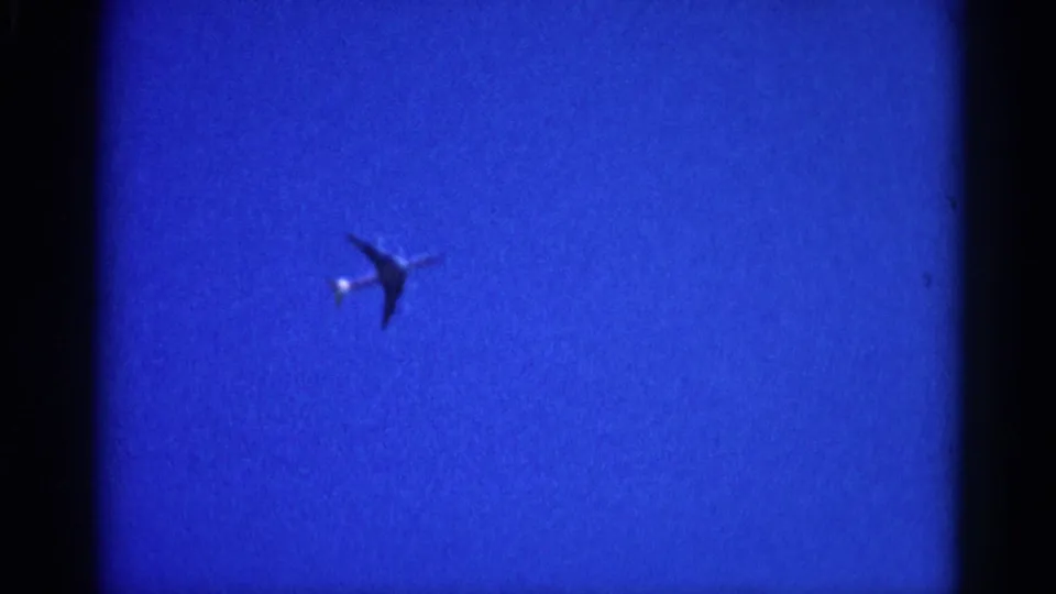 8mm home movie frame from 1962 Illinois showing a commercial jet as a dark silhouette against a uniformly deep blue sky, with a bright light near the nose.