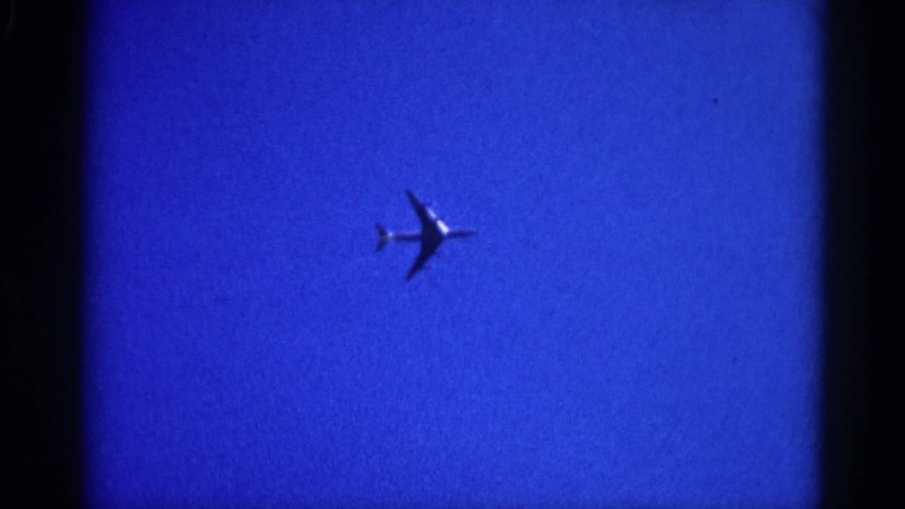 8mm film frame circa 1962 Illinois showing a commercial jet silhouette mid-crossing a blue sky; grain and soft focus are characteristic of the era and format.