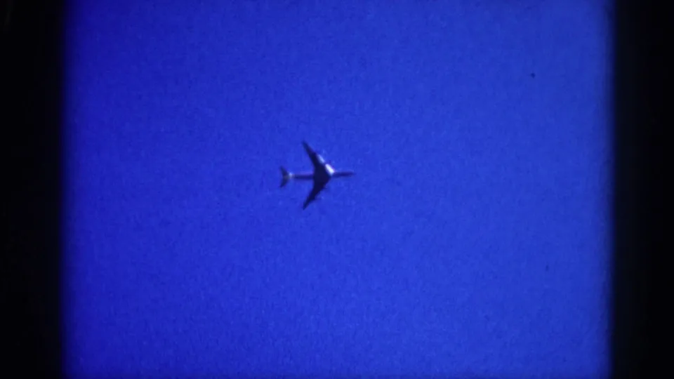 8mm film frame circa 1962 Illinois showing a commercial jet silhouette mid-crossing a blue sky; grain and soft focus are characteristic of the era and format.