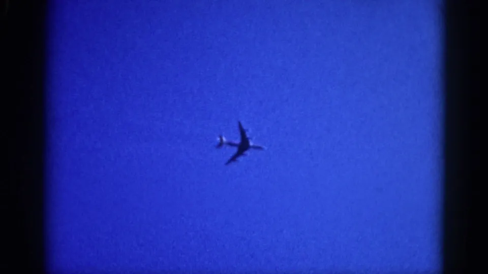 8mm home movie frame circa 1962 Illinois: jet aircraft receding into the lower portion of a uniformly blue sky, visibly smaller as it moves off-frame.
