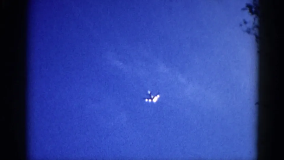 Final 8mm frame circa 1962 Illinois: the aircraft is visible only as clustered bright lights against a lighter dusk sky; dark foliage intrudes at the right frame edge.