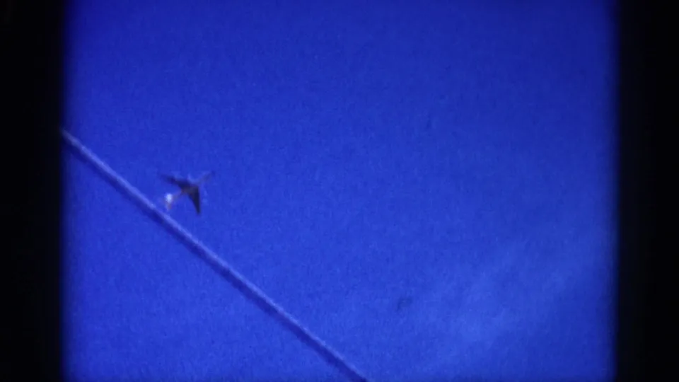 Small aircraft with bright light reflection passing beneath a diagonal overhead wire in 8mm-filmed blue sky, circa 1962 Illinois.