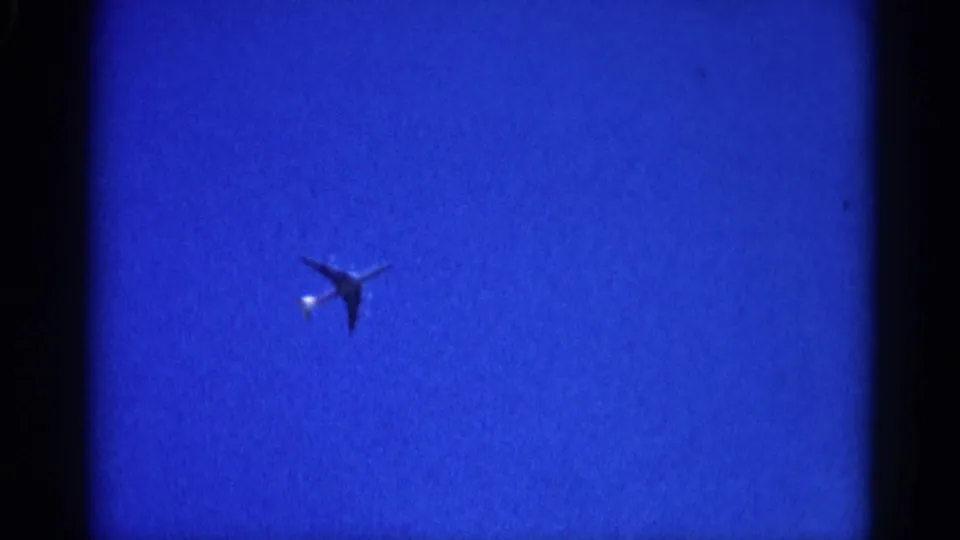 Small aircraft with bright fuselage or propeller reflection crossing a clear 8mm-filmed blue sky, circa 1962 Illinois.