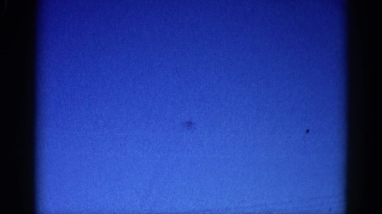 Tiny jet silhouette crossing a deep blue sky, captured on 8mm film in Illinois circa 1962; aircraft barely discernible through heavy grain.