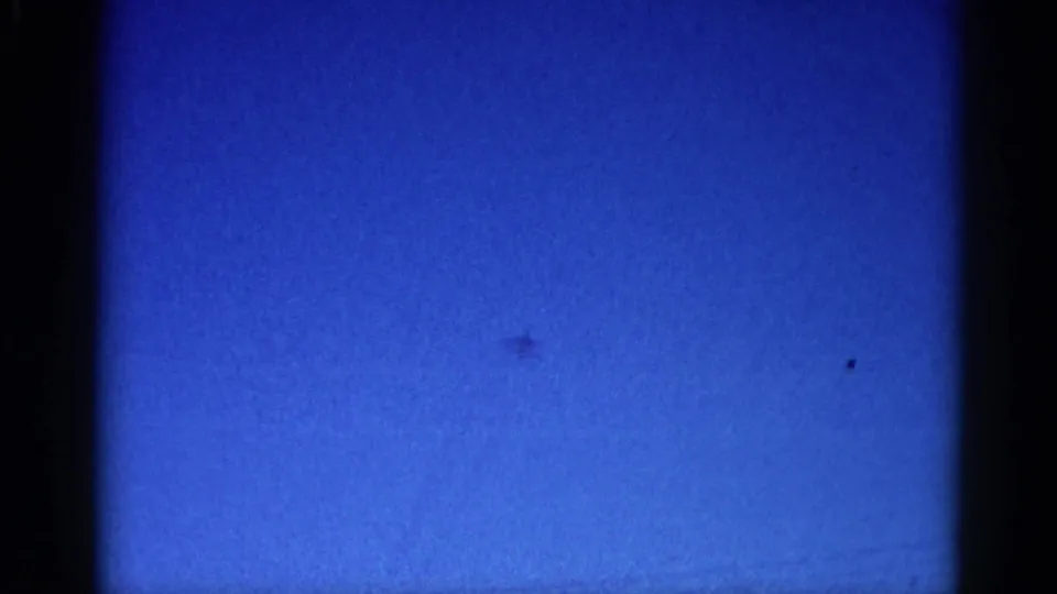 Tiny jet silhouette crossing a deep blue sky, captured on 8mm film in Illinois circa 1962; aircraft barely discernible through heavy grain.