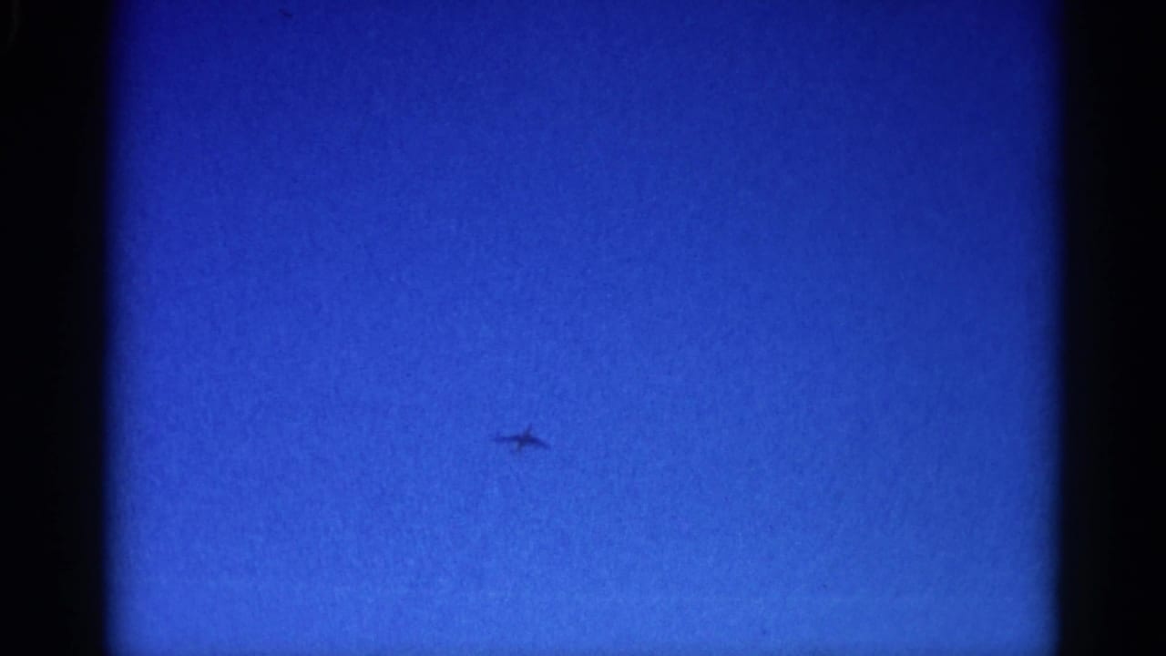 Jet silhouette with swept-wing profile crossing open blue sky on 8mm home-movie film, Illinois circa 1962.