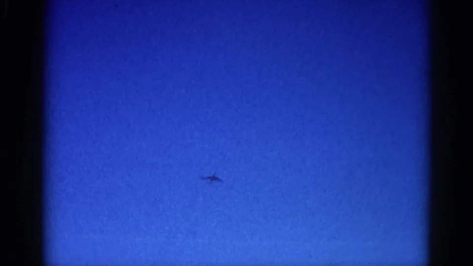 Jet silhouette with swept-wing profile crossing open blue sky on 8mm home-movie film, Illinois circa 1962.