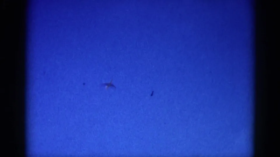 Jet silhouette near center-right with a second small mark nearby, filmed on 8mm in Illinois circa 1962; second mark may be artifact or second aircraft.