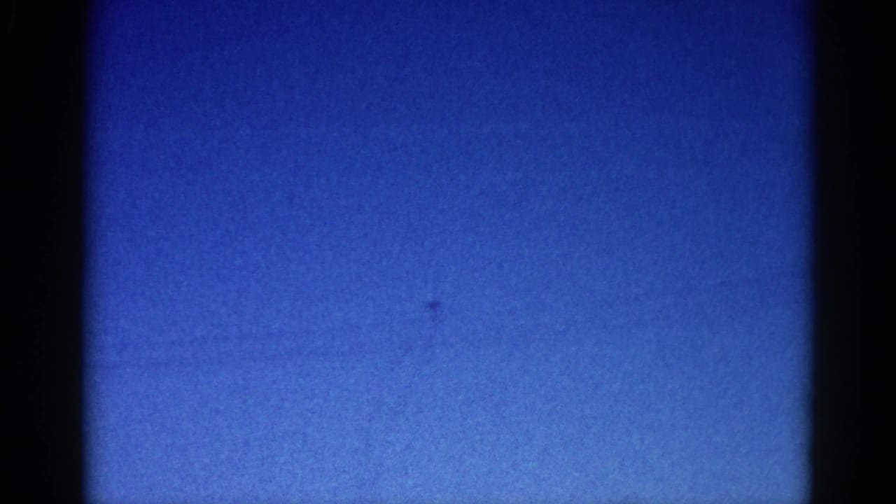 8mm home movie frame, circa 1962 Illinois: nearly featureless bright blue sky with a barely-visible distant aircraft speck and visible film grain.