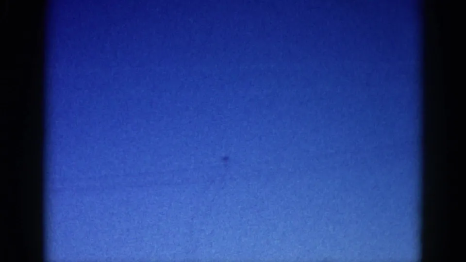 8mm home movie frame, circa 1962 Illinois: nearly featureless bright blue sky with a barely-visible distant aircraft speck and visible film grain.