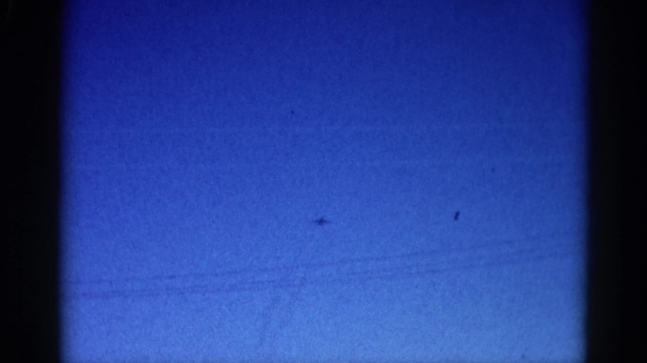 Blue sky with a tiny aircraft speck and faint diagonal linear marks across the frame; film grain visible throughout.