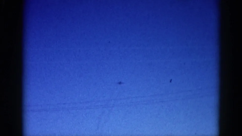 8mm home movie frame, circa 1962 Illinois: blue sky with a barely-visible aircraft and faint diagonal marks that may be contrails or film scratches.
