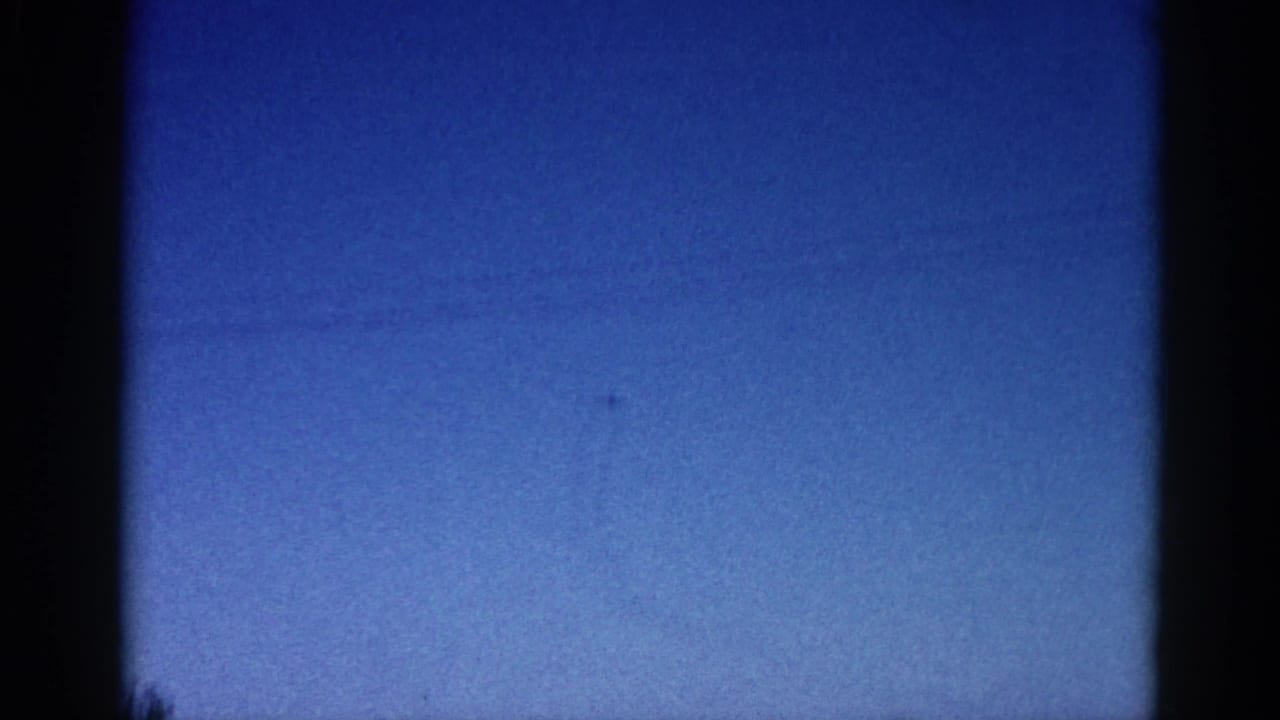 8mm home-movie frame circa 1962, Illinois: deep blue dusk sky with a small aircraft silhouette, visible film grain, and dark vignette framing.