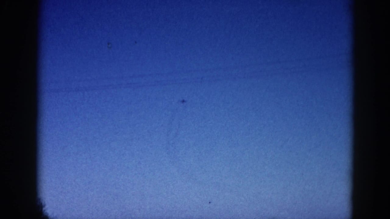 8mm archival frame circa 1962 showing a small aircraft traversing a deep blue twilight sky with heavy film grain and edge vignetting.