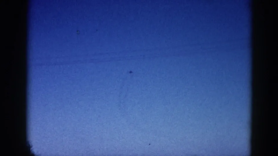 8mm archival frame circa 1962 showing a small aircraft traversing a deep blue twilight sky with heavy film grain and edge vignetting.